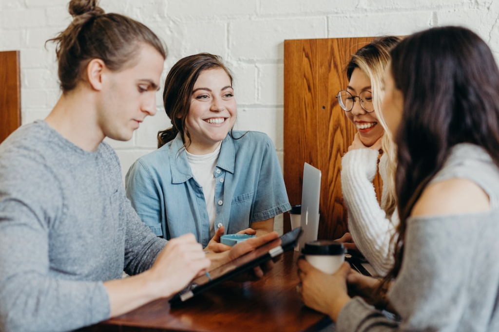 Four young adults sit at a wooden table engaged in discussion. One man uses a tablet, while the three women smile and converse. All are casually dressed, creating a relaxed atmosphere. Coffee cups and a laptop are visible on the table.