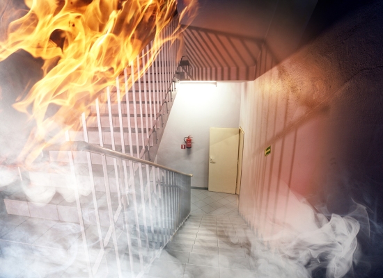 An emergency stairwell filled with smoke and flames. The metal stairs and white railing contrast with the light-colored walls. A fire extinguisher is mounted on the wall next to an open door, highlighting fire door identification in the UK, while a green emergency exit sign is visible.