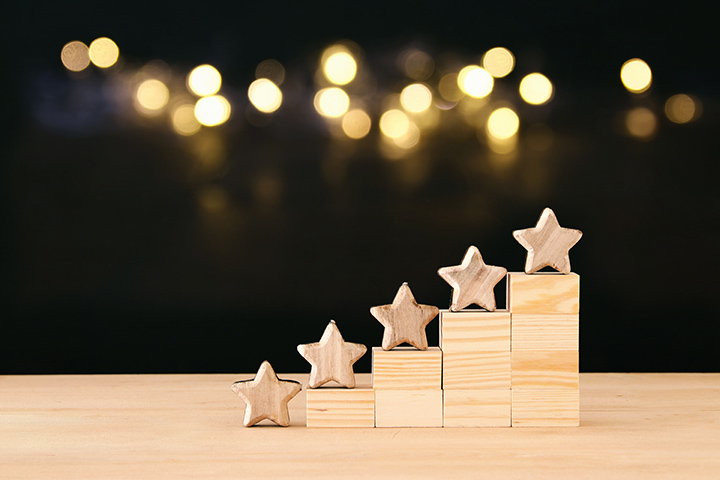 Wooden stars mounted on blocks of increasing height form a staircase pattern against a dark background with blurred golden lights. The arrangement suggests progress or achievement, much like how can I increase my Google 5-star reviews to highlight the stars more prominently as they ascend.