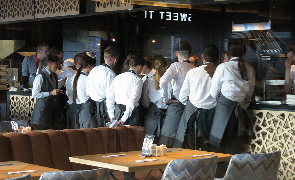 A group of restaurant staff wearing white shirts and gray aprons stand in a semi-circle, facing a counter and receiving instructions on how to train your staff to ask for more reviews from someone out of the shot. The restaurant boasts modern decor with patterned wall panels and plush seating.