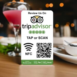 A table tent sign at a restaurant encourages patrons to review their experience on TripAdvisor Tap or Scan Smart Review Cards - Free Delivery. The sign, featuring the familiar logo, directs customers to "TAP or SCAN" using the NFC symbol and QR code. A cocktail and food plates enhance the inviting background ambiance.