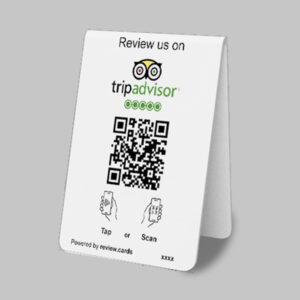 A small, white table tent displays a "Review us on TripAdvisor" message with the TripAdvisor logo. It includes a black QR code in the center, with images of hands tapping or scanning a phone. Text at the bottom reads, "Powered by review.cards.