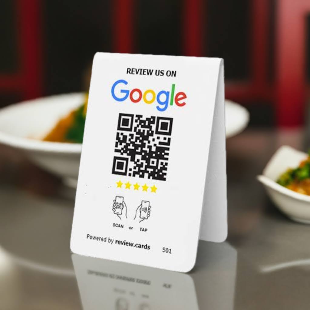 The Hospitality Bundle 1 table tent sign encourages customers to review a service on Google and TripAdvisor. It features a QR code, star rating graphic, and icons indicating "Scan" or "Tap." The bottom text reads "Powered by review.cards," with plates of food blurred in the background.