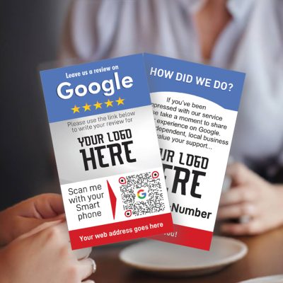 Two people are seated at a table holding Google review cards in the UK. The cards feature QR codes for easy access, a section for the business's logo, and prompts to leave a review by scanning with a smartphone. Text space is provided for a custom website address.